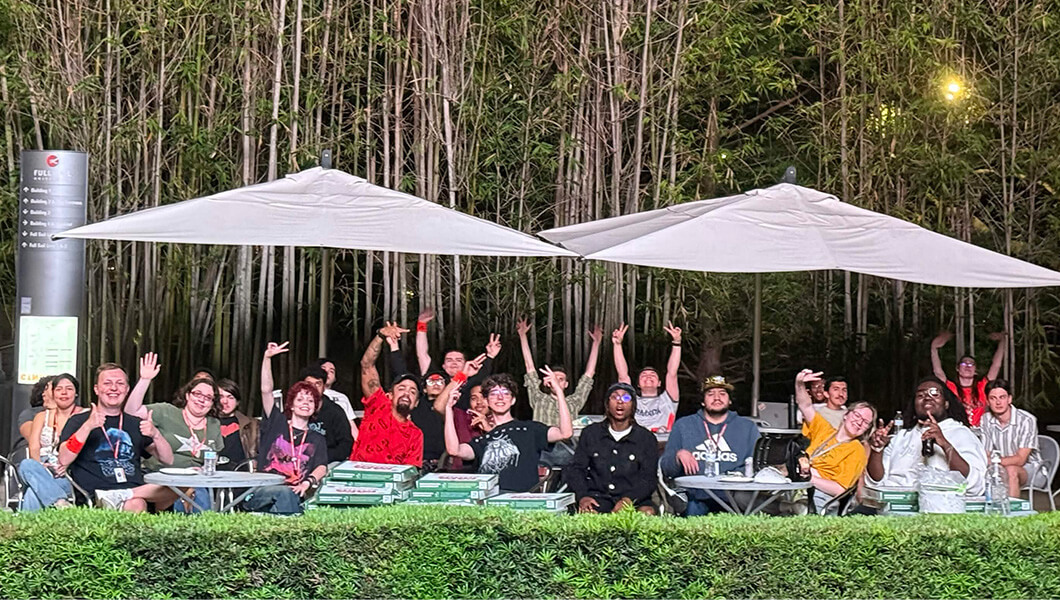 Una multitud de estudiantes saluda a la cámara mientras se sientan en mesas bajo sombrillas con una pila de cajas de pizza cerca y árboles detrás de ellos.